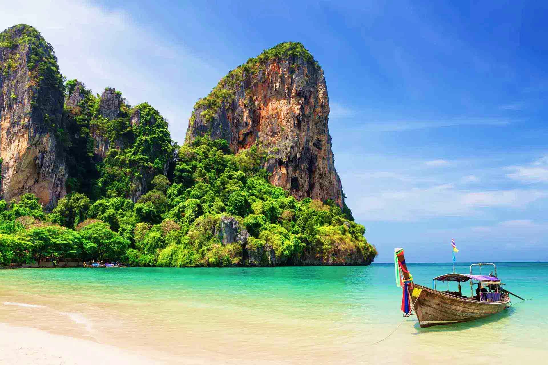 Limestone cliffs covered in green vegetation rising from turquoise water with traditional longtail boat on beach in Thailand